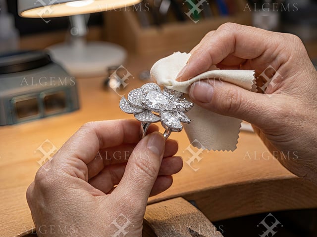 Two hands holding the Diamond Petal Ring by its band and gently wiping an upper white-metal edge with an ivory cloth at a wooden bench