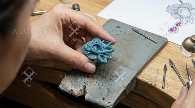 Blue-green floral wax ring model held over a worn grey bench support, with empty pear-shaped settings and tools nearby